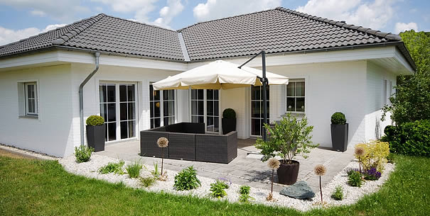 Ein weißer Eckbungalow mit schwarzem Dach und einer Terrase mit anliegendem Garten