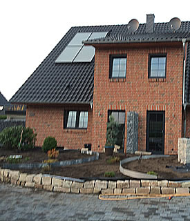 Der Eingagsbereich eines mit roten Verblender versehenes Doppelhauses. Im Verdergrund ist eine neu angelegter Garten mit einer Natursteinmauer